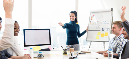 Réunion animée autour d'une table, les participants sourient, plusieurs lèvent la mains, et l'animatrice pointe du doigt pour donner la parole